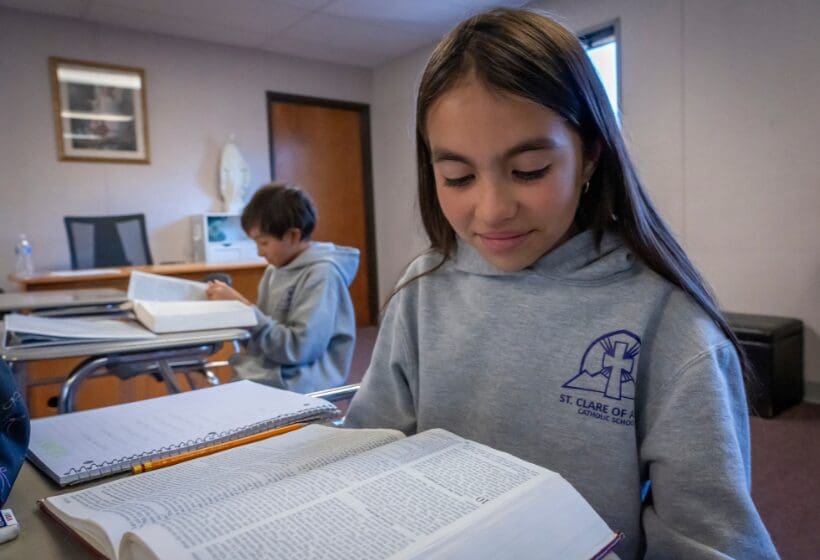 A girl reading a book in a classroom with another student in the background.