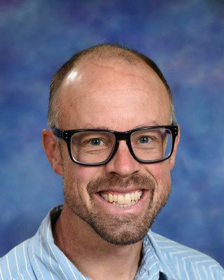 Smiling man with glasses and blue shirt against a blue background.