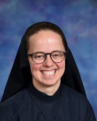 A smiling nun wearing glasses and a black habit against a blue background.