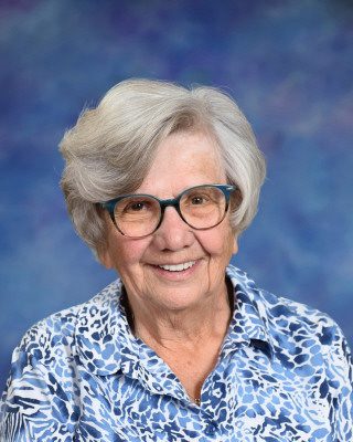Elderly woman smiling wearing glasses and a blue floral shirt.