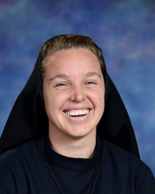 A smiling woman wearing a black religious habit against a blue background.