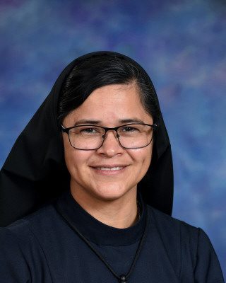 Portrait of a smiling nun wearing glasses and a black habit against a blue background.