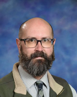 Portrait of a bald man with glasses and a beard wearing a suit jacket and tie.