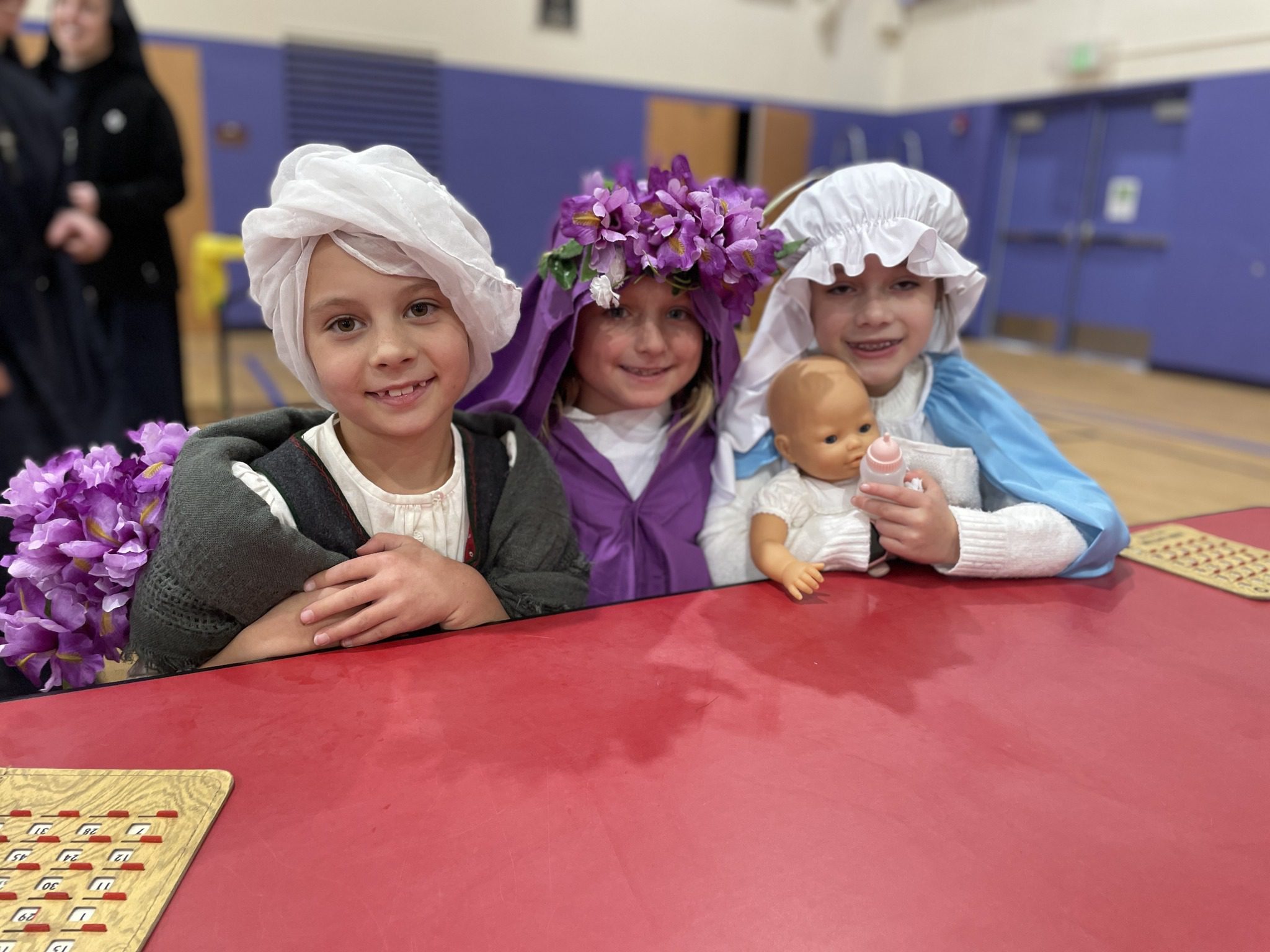 Three children dressed in historical costumes sitting at a red table, smiling.