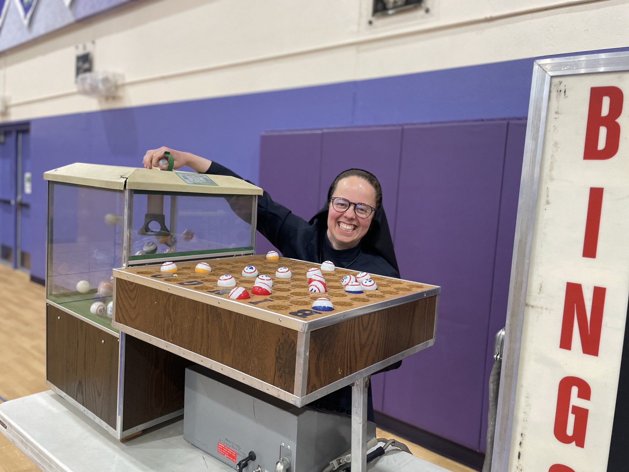 A smiling nun leaning on a display case with cupcakes.