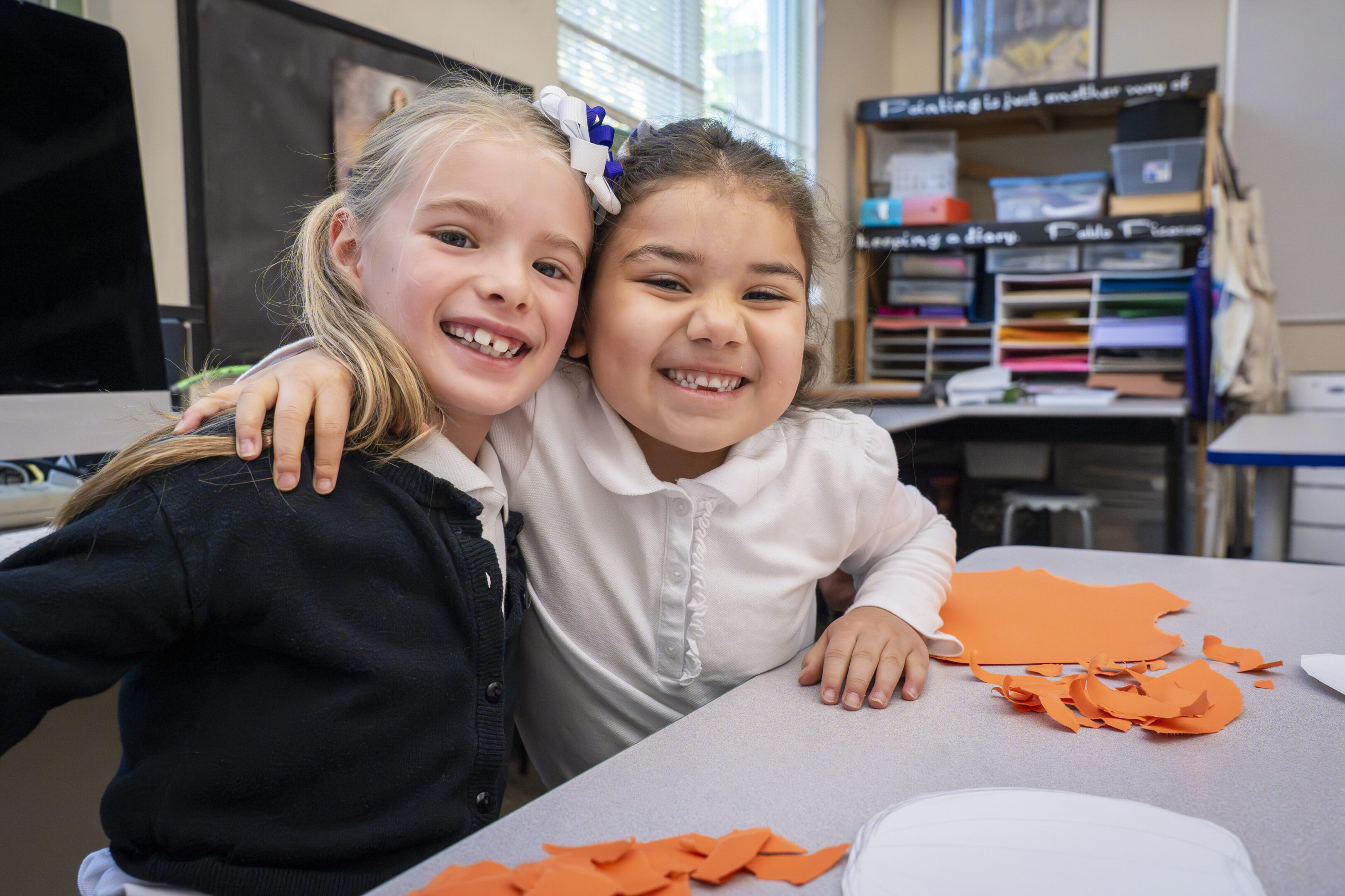 Two young girls smiling and hugging at a classroom table.