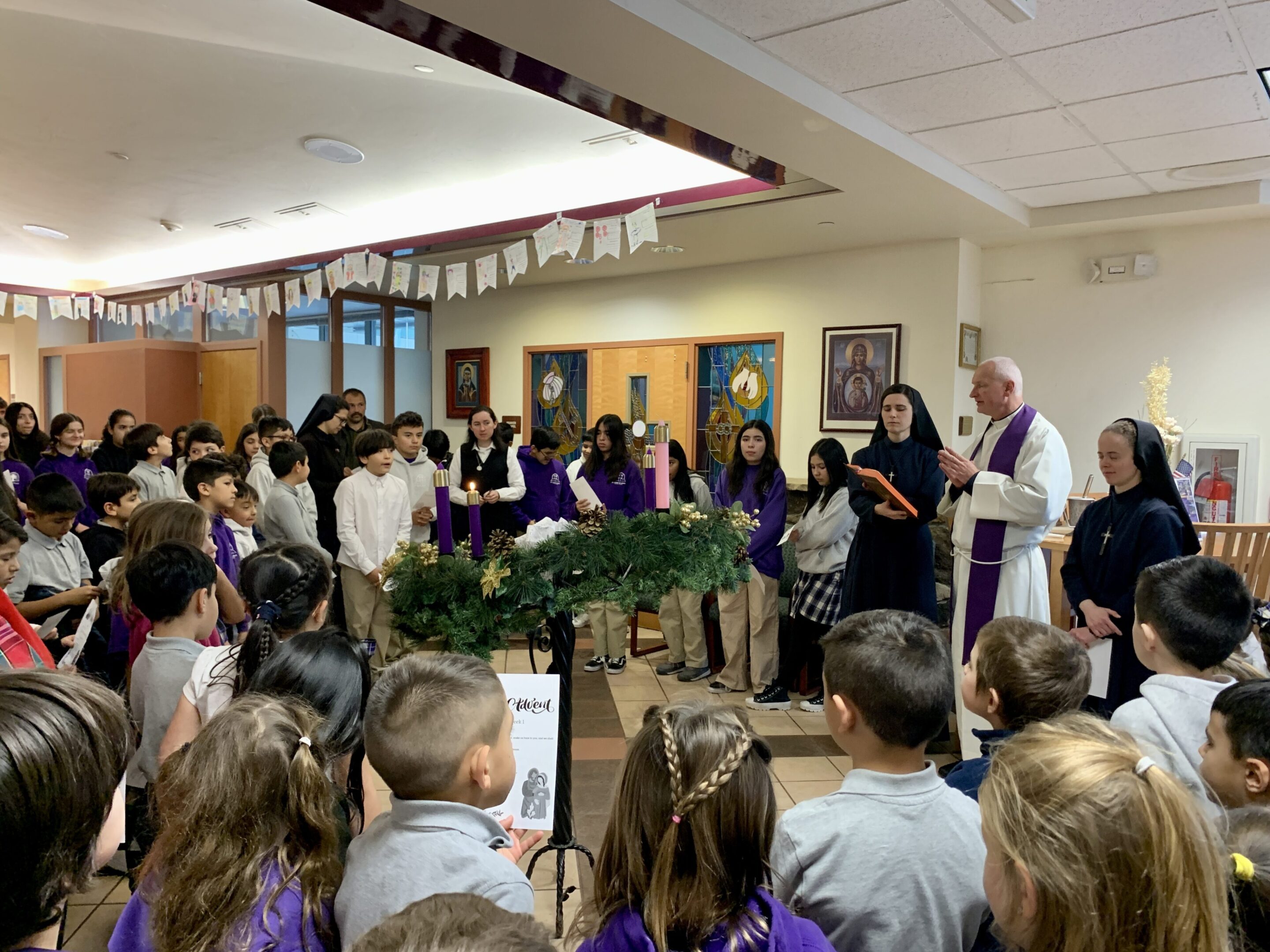 A group of children and adults gathered for a festive event with a priest speaking.
