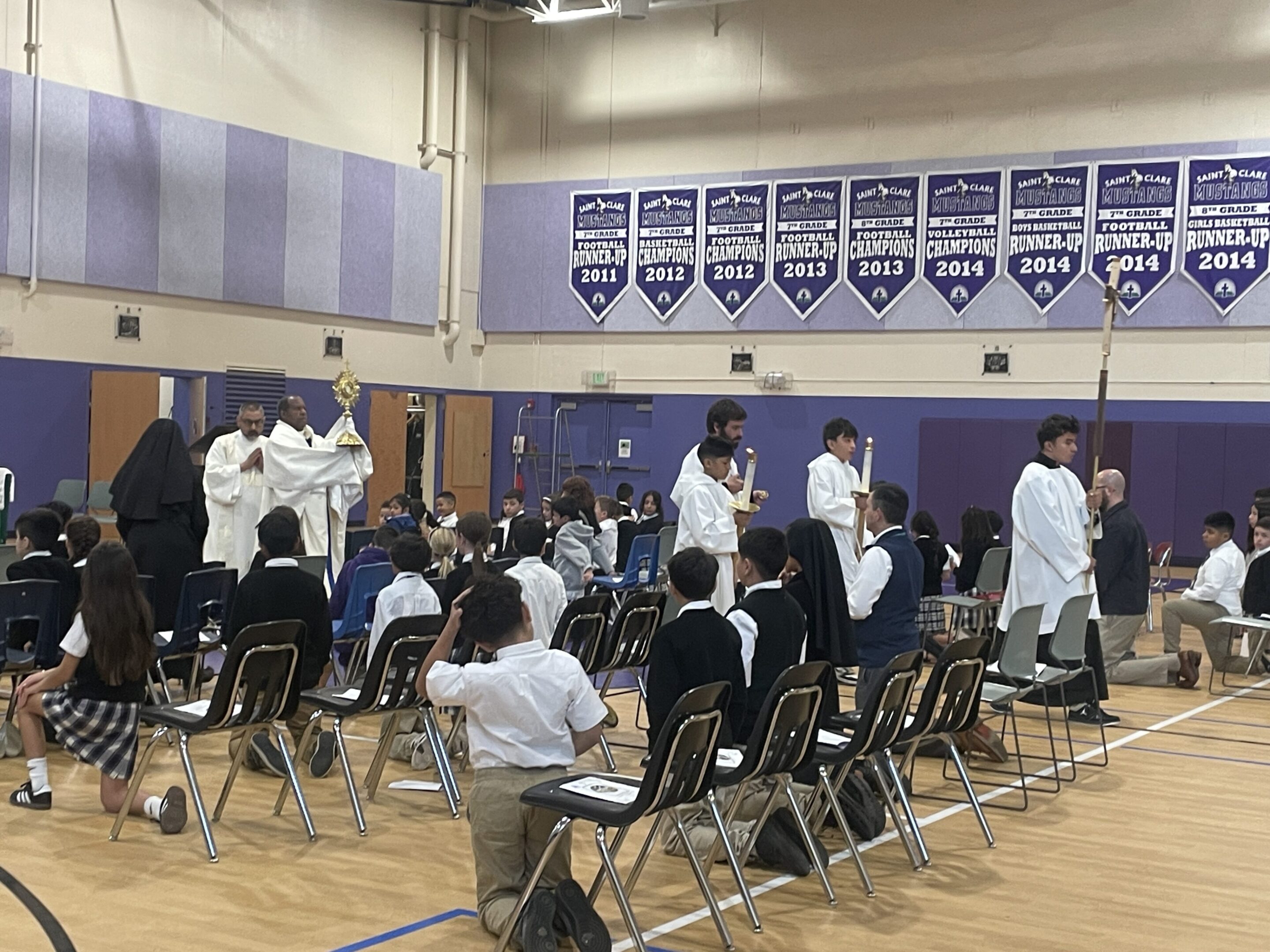 Students in white coats gather in a school gymnasium for an event.