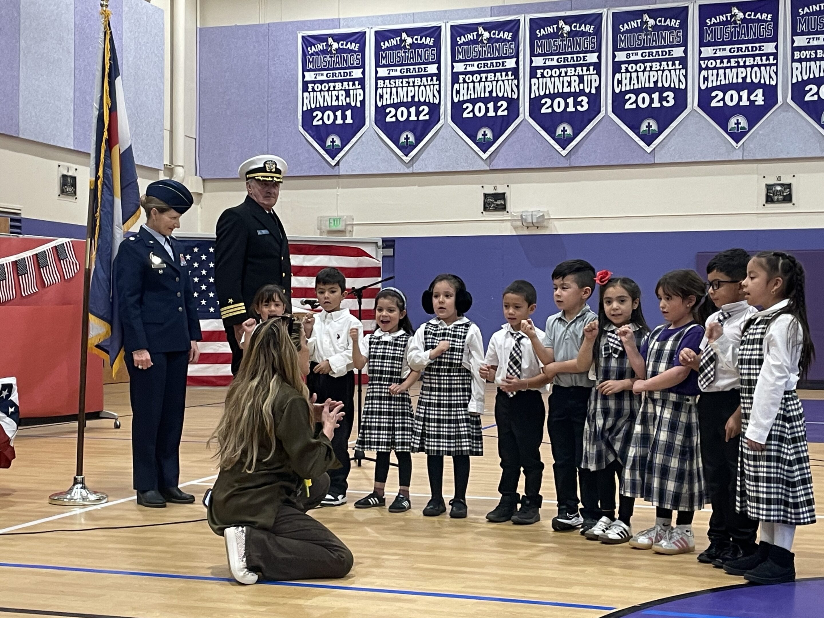 Children performing in a school assembly with a teacher and a guest in uniform.
