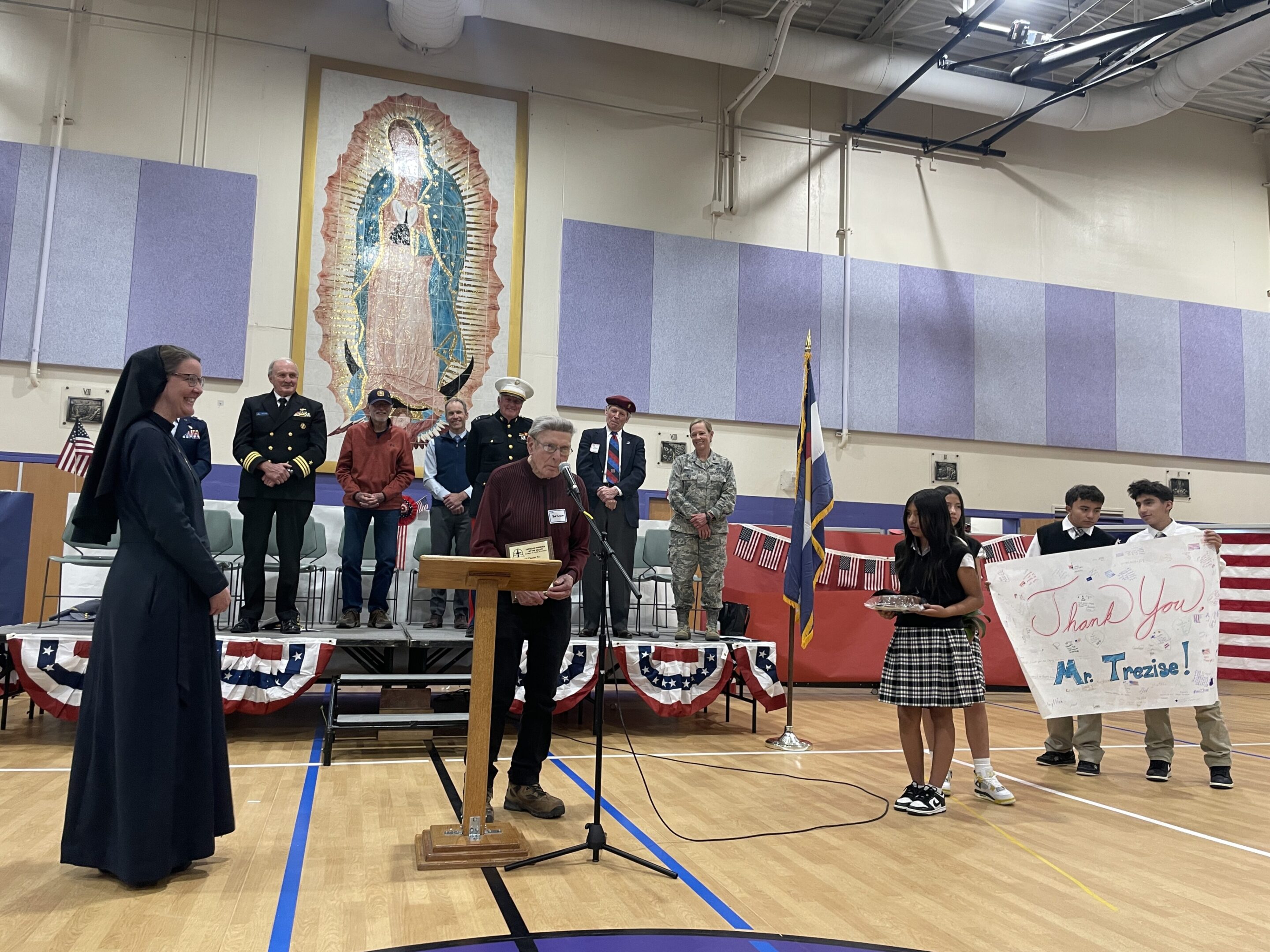 A group of people gathered in a gymnasium with a large religious painting on the wall.
