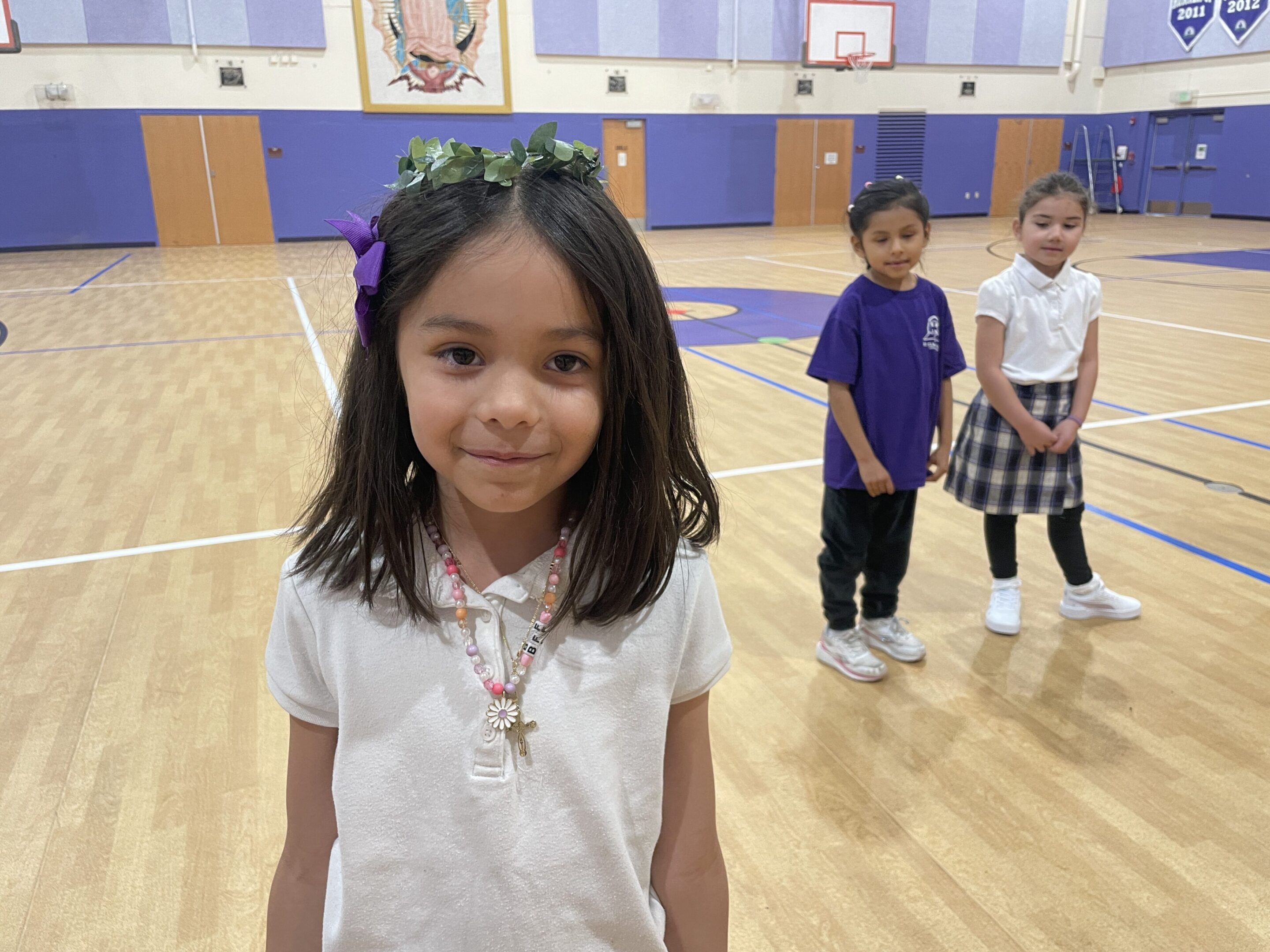 A young girl smiling in a gymnasium with two children in the background.