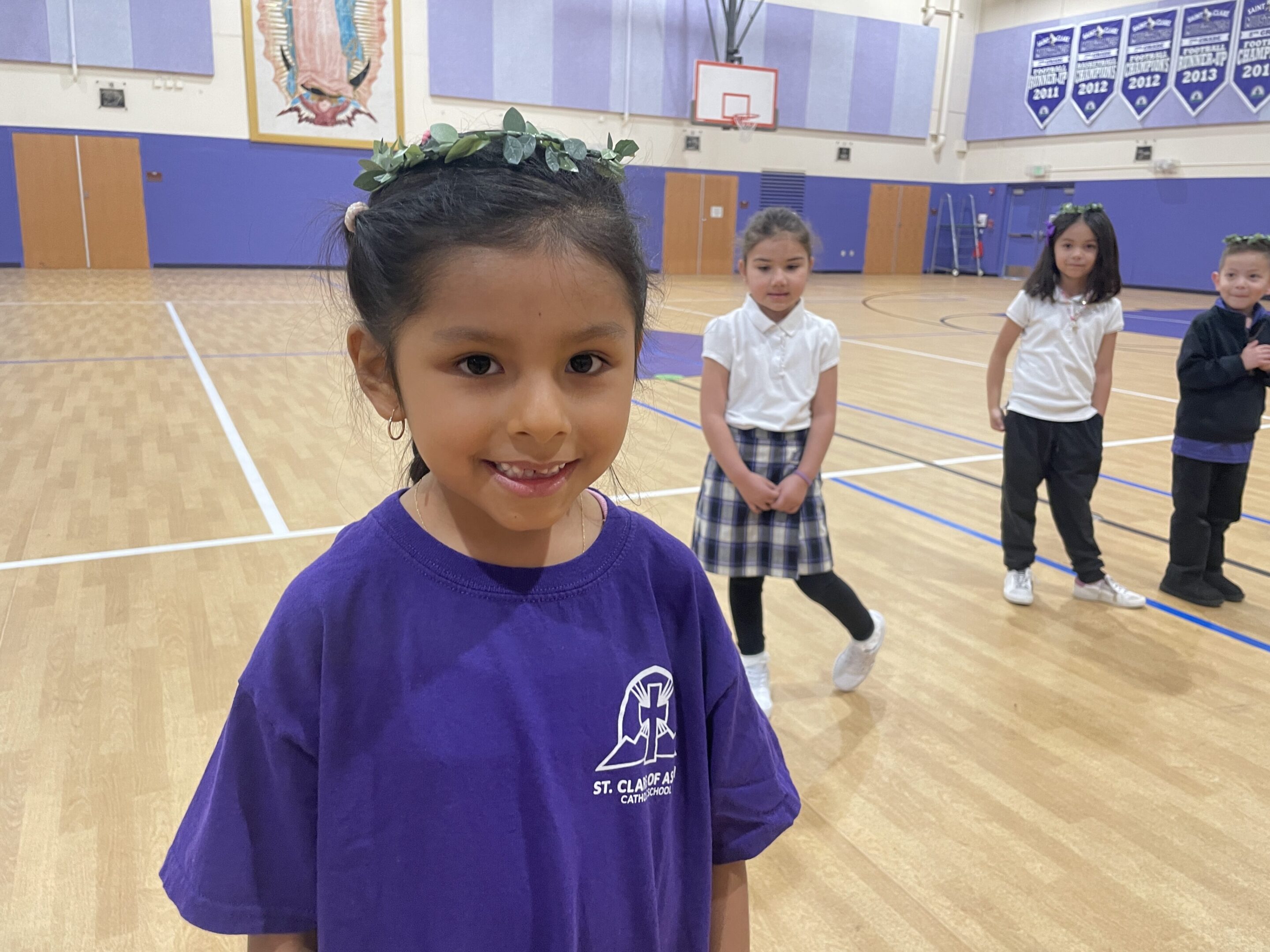 A smiling girl in a purple shirt stands in a gym with other children behind her.