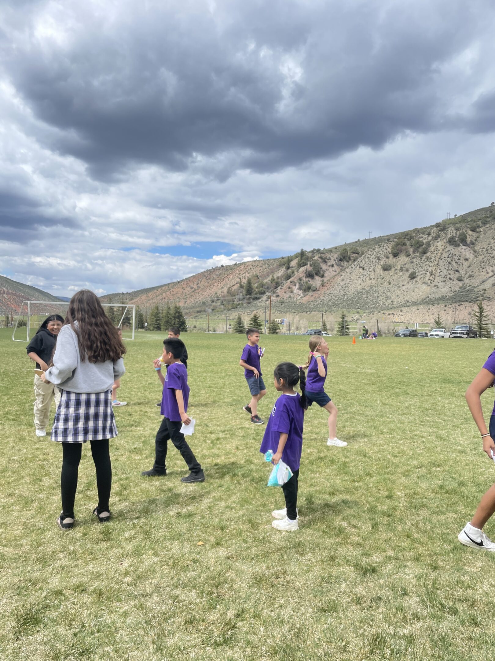 Children playing soccer on a field with adults supervising under a cloudy sky.