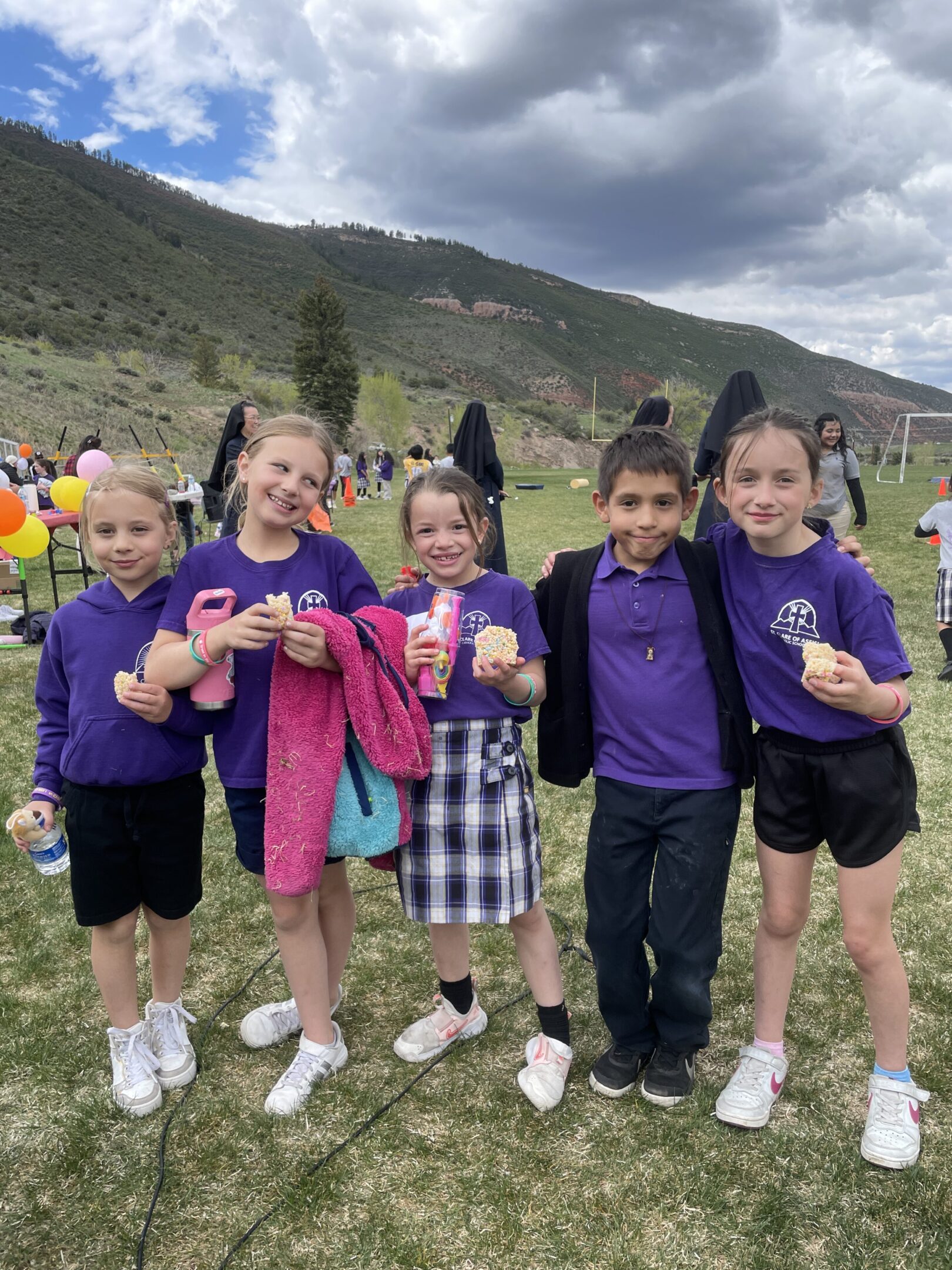 Five children in purple shirts enjoying outdoor snacks together on a grassy field.