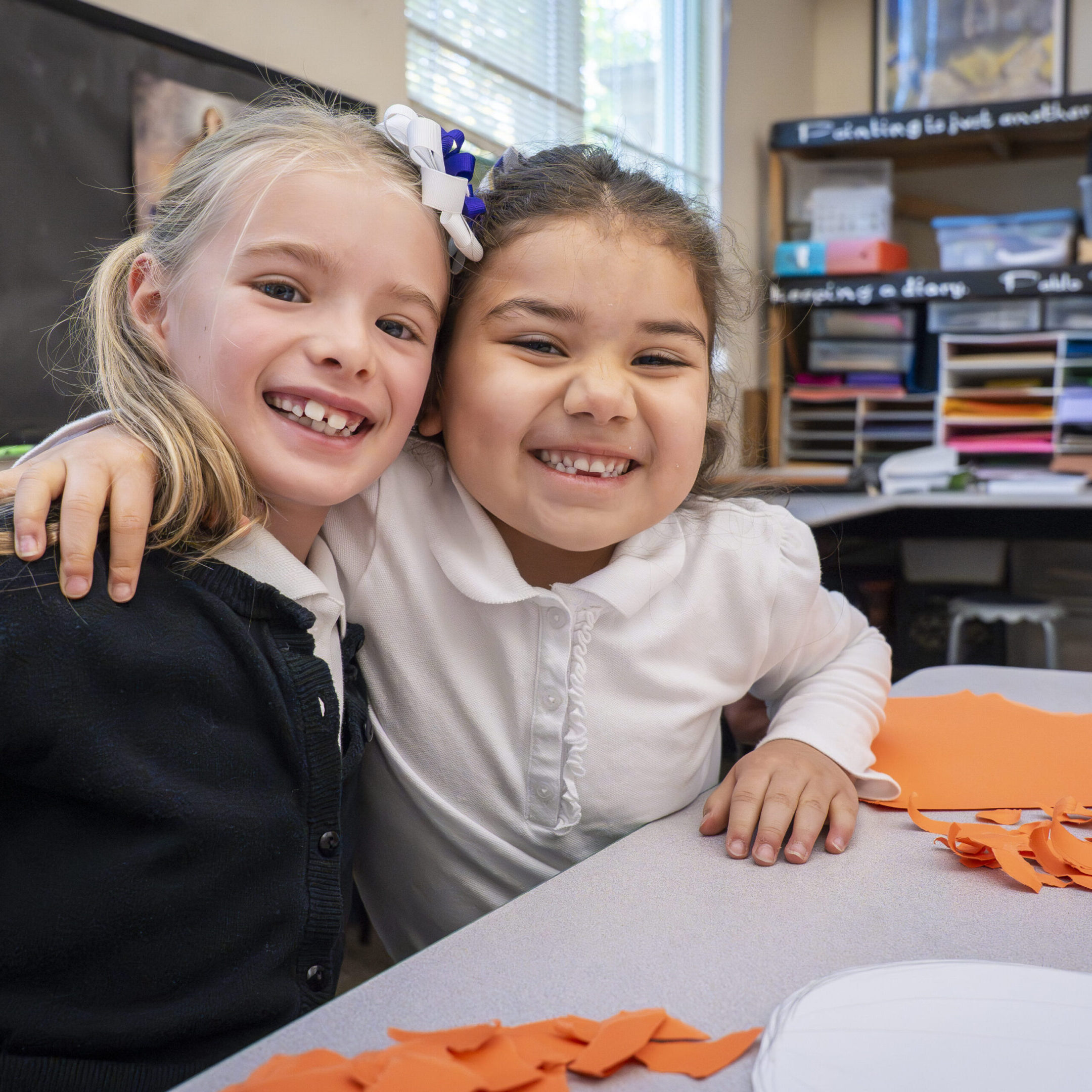 Two young girls smiling and hugging at a classroom table.