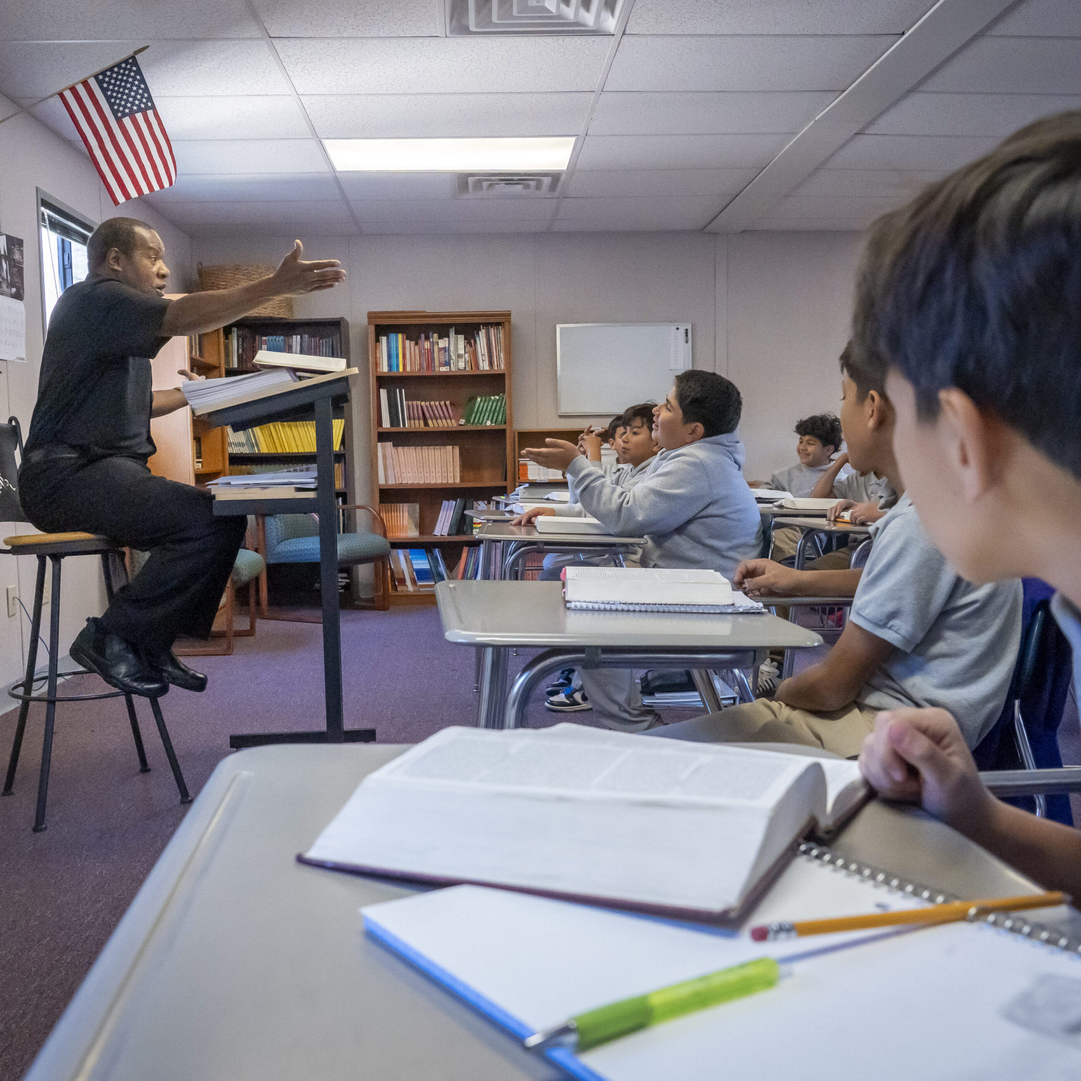 Teacher instructing students in a classroom with books and notebooks.